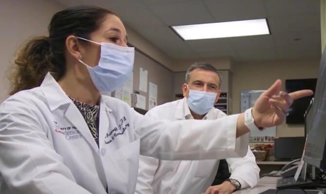 A man and woman in UT MD Anderson lab coats wearing face masks. The woman is pointing to a computer screen.