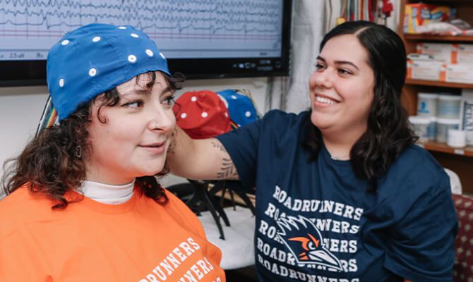 Two smiling women in UTSA Roadrunner shirts sit together. One wears a neurofeedback cap.