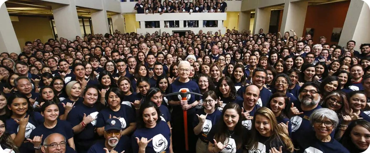 Diana Natalicio, in the middle of the hall, holding a pick-axe, surrounded by students and staff in UT El Paso colors and wear.