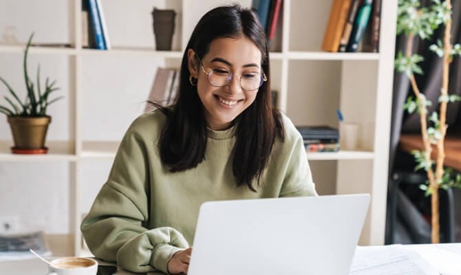 Person working at a laptop in a home office with shelves and plants in the background.