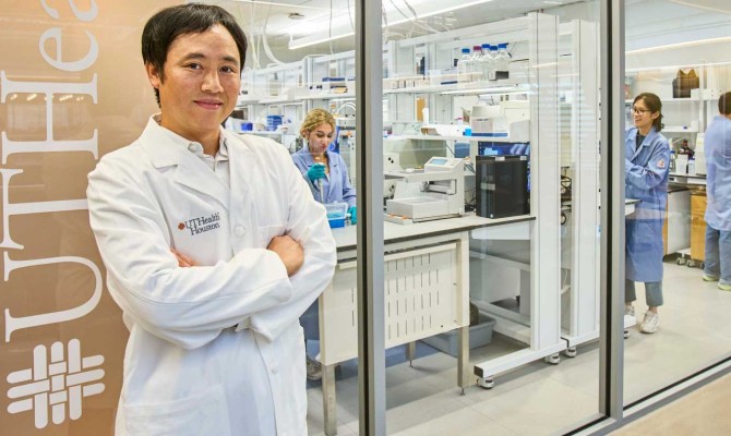 Kyoji Tsuchikama, PhD, standing in front of a lab wearing a white coat and folding his arms. 