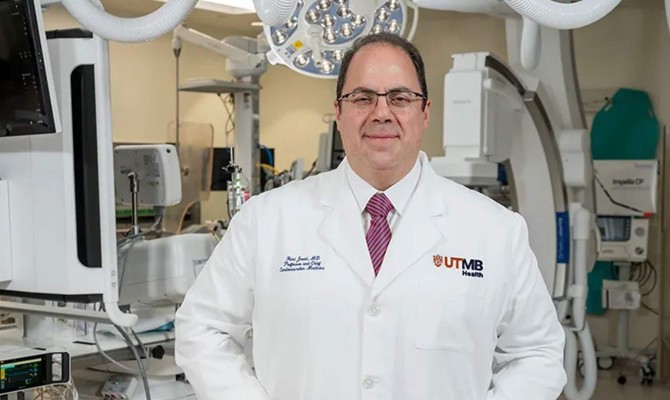 Clinician wearing a white lab coat with the UTMB Health logo stands in a cardiac procedure room with imaging equipment and monitors visible behind them.