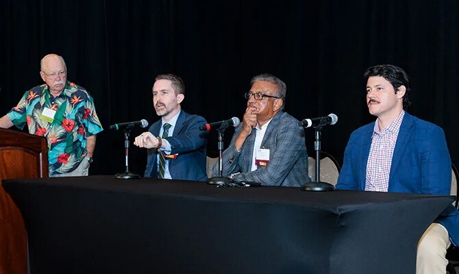 Three men in business attire sit at a long black table while a man in a colorful, tropical shirt stands at a podium during a panel discussion.