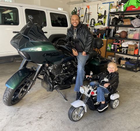 Derek Horton, UT System's assistant vice chancellor, Office of Budget and Planning, sitting on his Harley Davidson motorcycle with his son sitting on a play motorcycle next to him in their garage