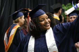 African American female wearing a cap and gown and holding a diploma with other graduating students in the background.